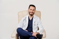 Arabian doctor surgeon in medical robe with phonendoscope seats and smile in armchair in studio on white blanked