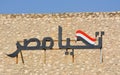 Arabic calligraphy text Tahya Misr (Long live Egypt) with the Egyptian flag on a hill with a blue sky
