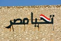 Arabic calligraphy text Tahya Misr (Long live Egypt) with the Egyptian flag on a hill with a blue sky