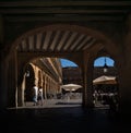 Arched entrance with a wooden roof to the main square of Salamanca with tourists walking, umbrellas and terraces of bars and the