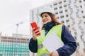 Happy Architect with a blueprints using smartphone at a construction site. Portrait of redhead woman constructor wearing