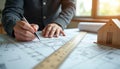 Architect draws architectural project with scale ruler. Model house stands on blueprint. Person sketches construction plan for
