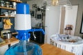 Arger bottle of clean water 19 liters with blue pomp in the interior of the apartment with a baby cot in the background. Clean and
