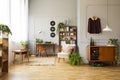 Armchair next to plant in vintage living room interior with wooden cabinet and chair at desk. Real photo