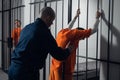 An armed guard searches a newly arrived criminal in a prison corridor against a backdrop of bars.