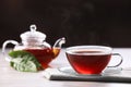 Aromatic hot tea in glass cup, teapot and leaves on white wooden table against black background. Space for text Royalty Free Stock Photo