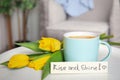 Aromatic morning coffee, beautiful flowers and card with RISE AND SHINE wish on white table indoors