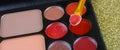 An array of makeup or cosmetics scattered over a gold glitter background. A sponge applicator is loaded with red lip color.