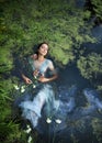 Art beautiful romantic woman lies in swamp in blue long dress with flowers. Portrait brunette in transparent dress in water swamp