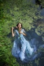 Art beautiful romantic woman lies in swamp in blue long dress with flowers. Portrait brunette in transparent dress in water swamp Royalty Free Stock Photo