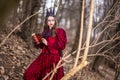 Art Photography. Mysterious Fairy Lady in Red Dress and Black Crown With Old Book. Posing in Forest Outdoors