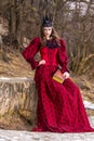 Art Photography. Mysterious Medieval Queen in Red Dress and Spiky Black Crown Posing With Ancient Book in Forest in Early Spring