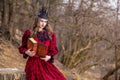 Art Photography. Mysterious Medieval Queen in Red Dress and Spiky Black Crown Posing With Ancient Book in Forest in Early Spring