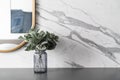 Artificial plant in glass vase compose with gold stainless mirror frame on gray spray-painted working table with marble wall in