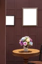 Artificial rose flowers in ceramic vase on wooden round table with teak wood chair and the old blank picture frame on wooden wall Royalty Free Stock Photo