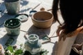 An artisan applies green paint to a canvas using a brush, surrounded by various natural materials and tools on a sunlit table in a