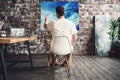 Artist in art studio sits on chair in front of canvas on easel and drawing. Painter work in workshop. Working process Royalty Free Stock Photo