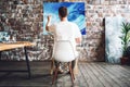 Artist is paint an oil painting in art studio while sitting on a chair in front of a canvas. Painter drawing process in loft works