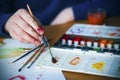 The artist`s hand, with soiled fingers, gracefully holds a paintbrush, mixing paints on a plastic palette, and a sheet of paper Royalty Free Stock Photo