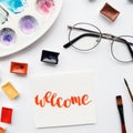 Artist workspace. Word Welcome written in calligraphy style, watercolor cuvettes and palette on a white background. Royalty Free Stock Photo