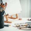 Artistic equipment in painter studio: easel, paint brushes, tubes of paint, palette and paintings on work table. Royalty Free Stock Photo