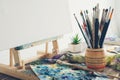 Artistic equipment in studio: canvas on wooden easel, paint brushes, paints and used palette. Royalty Free Stock Photo