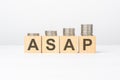 ASAP text written on wooden block with stacked coins on white background