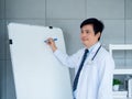 Asian adult male doctor in white uniform with stethoscope is holding chemical pen, writing on a blank whiteboard. Royalty Free Stock Photo