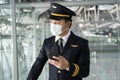 Asian Airliner pilot wearing face mask, standing in airport terminal. Young handsome captain using mobile phone chatting and