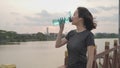 Asian athlete woman in green nature tree park and lake in evening sunset feeling thirsty drink water from bottle. energy drinks