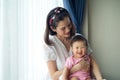 Asian beautiful mother holding her cute baby in her arms sitting near the window at home. Royalty Free Stock Photo