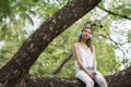 Asian Beautiful woman sit on big tree and listen to online streaming music from smartphone by earbuds. Brunette girl relax in Royalty Free Stock Photo