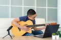 Asian boy playing acoustic guitar and watching online course on laptop while practicing at home. Asian boy virtual happy hour Royalty Free Stock Photo