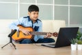 Asian boy playing acoustic guitar and watching online course on laptop while practicing at home. Asian boy virtual happy hour Royalty Free Stock Photo