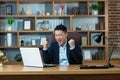 Asian businessman shouts with joy and holds hands up, celebrates victory, man works at desk in office, looks at laptop screen
