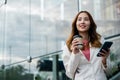 Asian businesswoman with smartphone and cup coffee standing against street building near office Royalty Free Stock Photo