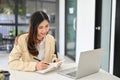 Asian businesswoman taking notes on her notebook, looking at laptop screen Royalty Free Stock Photo