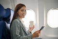 Asian businesswoman using smartphone and sipping coffee during flying to somewhere by plane