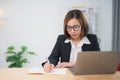 Asian businesswoman wearing suit and glasses writing notes book and working laptop on wooden table at home. Entrepreneur woman Royalty Free Stock Photo