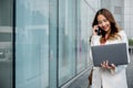 Asian businesswoman working on laptop and talking on cell phone at front building near office Royalty Free Stock Photo