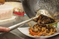 An Asian chef stir-fries caramelized meat in a steaming wok with a large ladle and pours it onto a plate to serve the diner