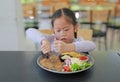 Asian child girl eating Pork steak and vegetable salad on the table with holding knife and fork. Children having breakfast Royalty Free Stock Photo