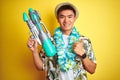 Asian chinese man wearing floral hawaian lei and water gun over isolated yellow background happy with big smile doing ok sign, Royalty Free Stock Photo