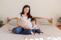 Asian Chinese pregnant woman with toddler girl sitting on bed at home. Girl daughter kid hugging mom belly. Mother and baby