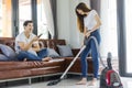 Asian couple girl doing floor cleaning with vaccuum cleaner while man on a brown sofa at home.