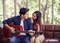 Asian couple sitting close together on couch in living room , a man playing guitar to his girlfriend and they are looking to each Royalty Free Stock Photo