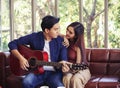 Asian couple sitting close together on couch in living room , a man playing guitar to his girlfriend and they are looking to each Royalty Free Stock Photo