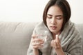 Asian Cute of  girl having headache and high temperature from illness holding pill of medicine and water sitting on sofa at home, Royalty Free Stock Photo