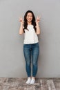 Asian cute woman in casual t-shirt and jeans smiling and gesturing victory sign with both hands, isolated over gray background