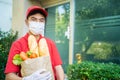 Asian deliver man wearing face mask in red uniform handling bag of food, fruit, vegetable waiting to give customer in front of the